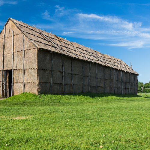 A Ganondagan Long House with green grass and a blue sky