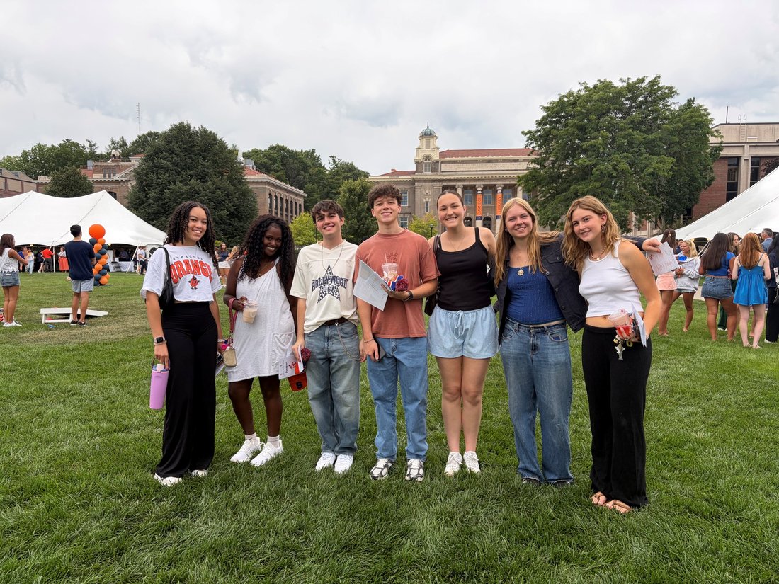 Seven students pose together on the quad during a campus event, with cornhole boards, orange and navy balloon columns, and Carnegie Library visible in the background.