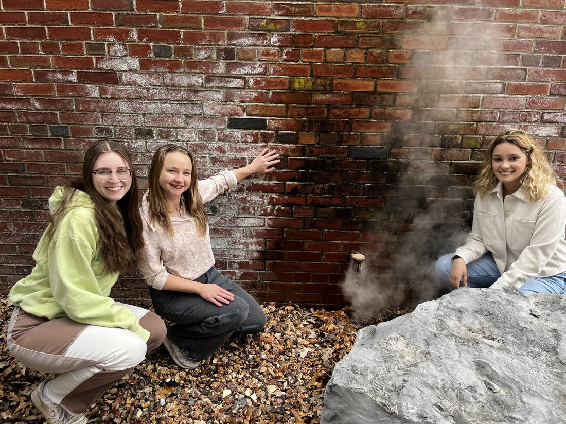 Three women bending down in front of a steam vent.