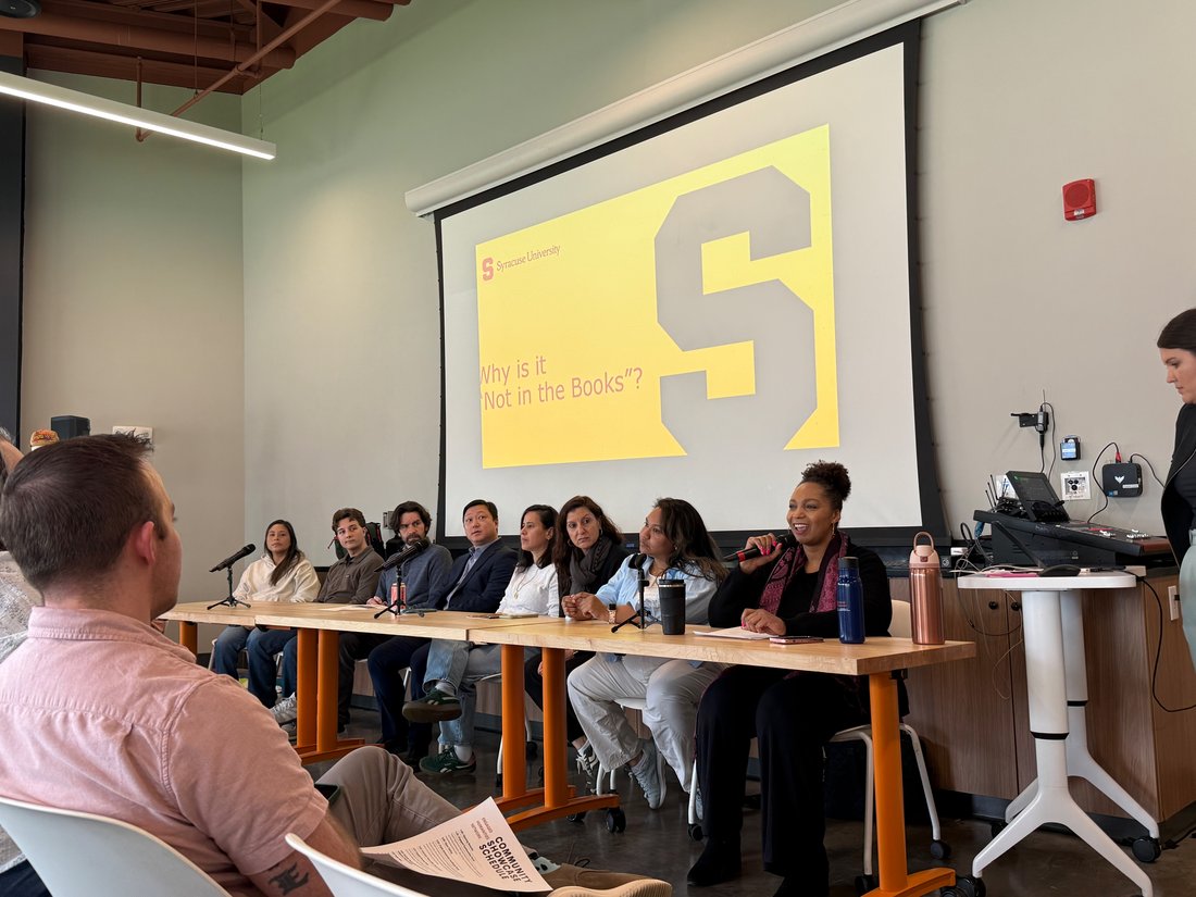 Eight panelists sit at the front of a Syracuse University classroom during a community-engaged learning event. One panelist speaks into a microphone while audience members listen. A projected slide behind the panel reads, “Why is it ‘Not in the Books’?”