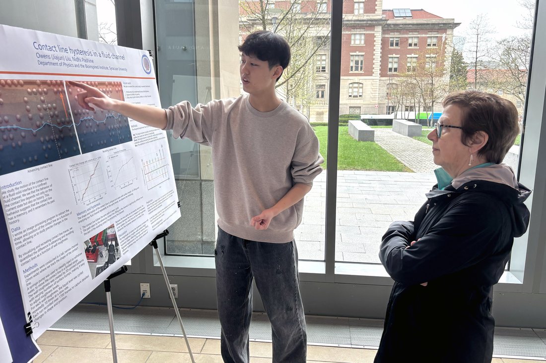 A student points to details on a research poster titled 'Contact Line Hysteresis in a Fluid Channel' while presenting his work to an attendee at an academic poster session.