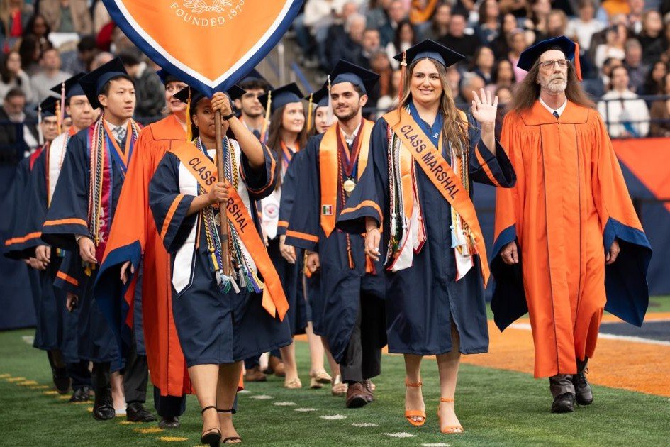 Graduates in caps and gowns process across a stadium floor during commencement, led by two Class Marshals wearing orange sashes and one carrying a Syracuse University banner.