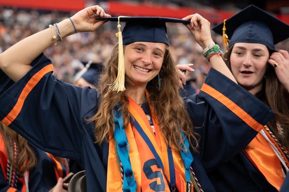 A smiling graduate adjusts her cap during a commencement ceremony, wearing an orange and blue Syracuse University stole over her gown.