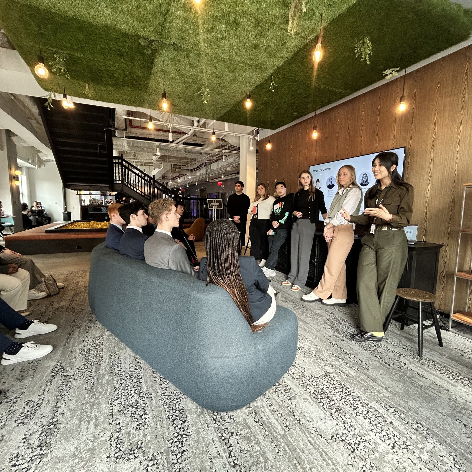A group of students attend an alumni panel event in a modern coworking space, with some seated on a curved gray sofa and others standing near a wall-mounted screen displaying a "Meet the Alumni" slide. The stylish space features wood-paneled walls, hanging Edison bulbs and a green moss ceiling.