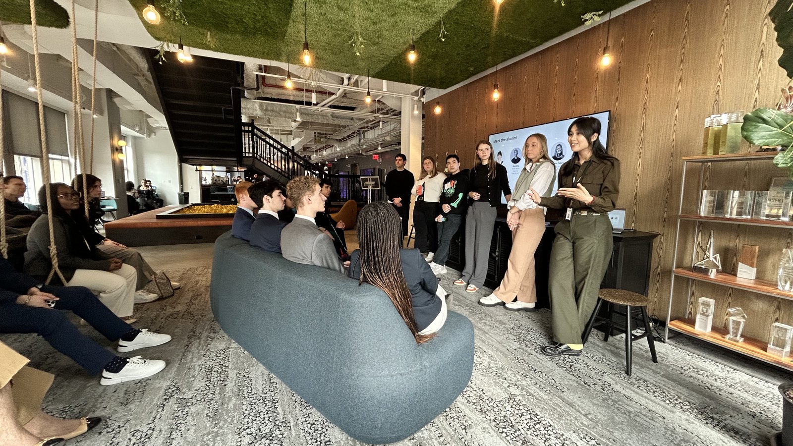 A group of students attend an alumni panel event in a modern coworking space, with some seated on a curved gray sofa and others standing near a wall-mounted screen displaying a "Meet the Alumni" slide. The stylish space features wood-paneled walls, hanging Edison bulbs and a green moss ceiling.