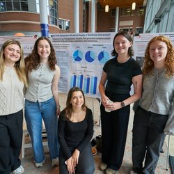 Five students smile in front of research poster boards at an academic symposium, with one poster titled 'Are First-Generation Students Happy at Syracuse University?' visible behind them.