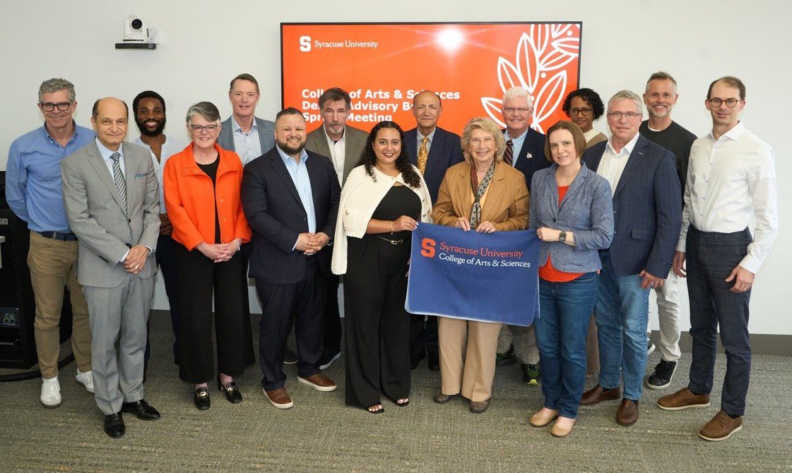 A group of 16 people stand side by side holding an A&S flag