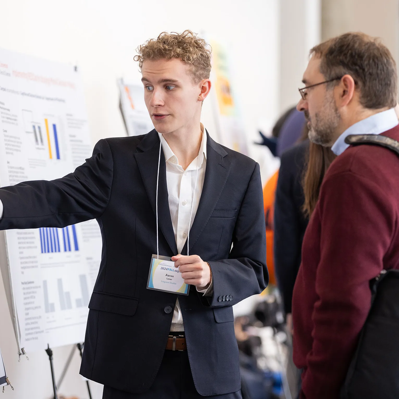 A young man in a dark suit gestures toward a research poster while presenting his work to an older man wearing a red sweater at an academic conference or research expo, with other attendees and posters visible in the background.