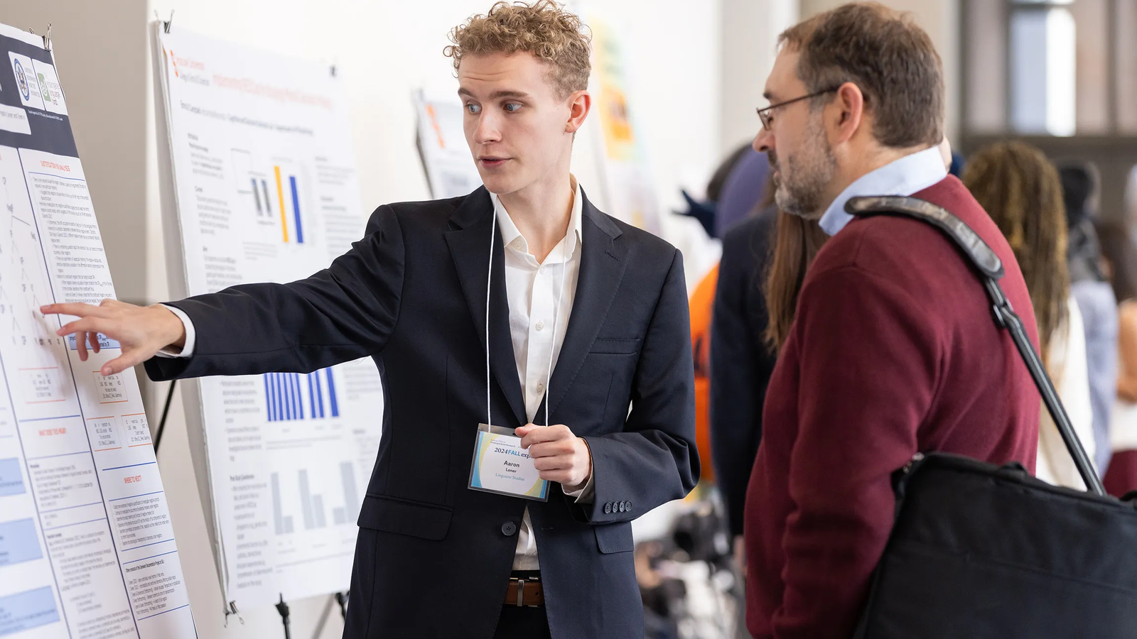 A young man in a dark suit gestures toward a research poster while presenting his work to an older man wearing a red sweater at an academic conference or research expo, with other attendees and posters visible in the background.