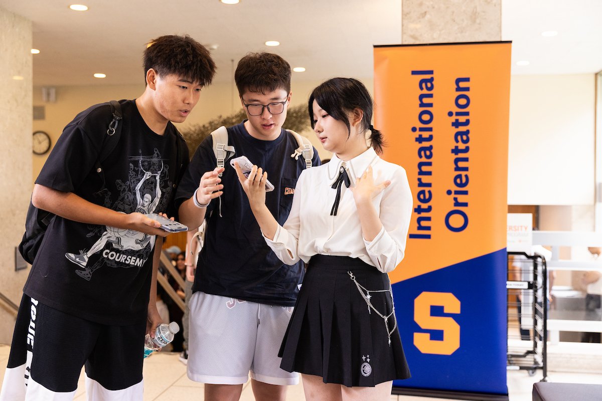 Three students look at a cell phone in front of a sign that reads "International Orientation."