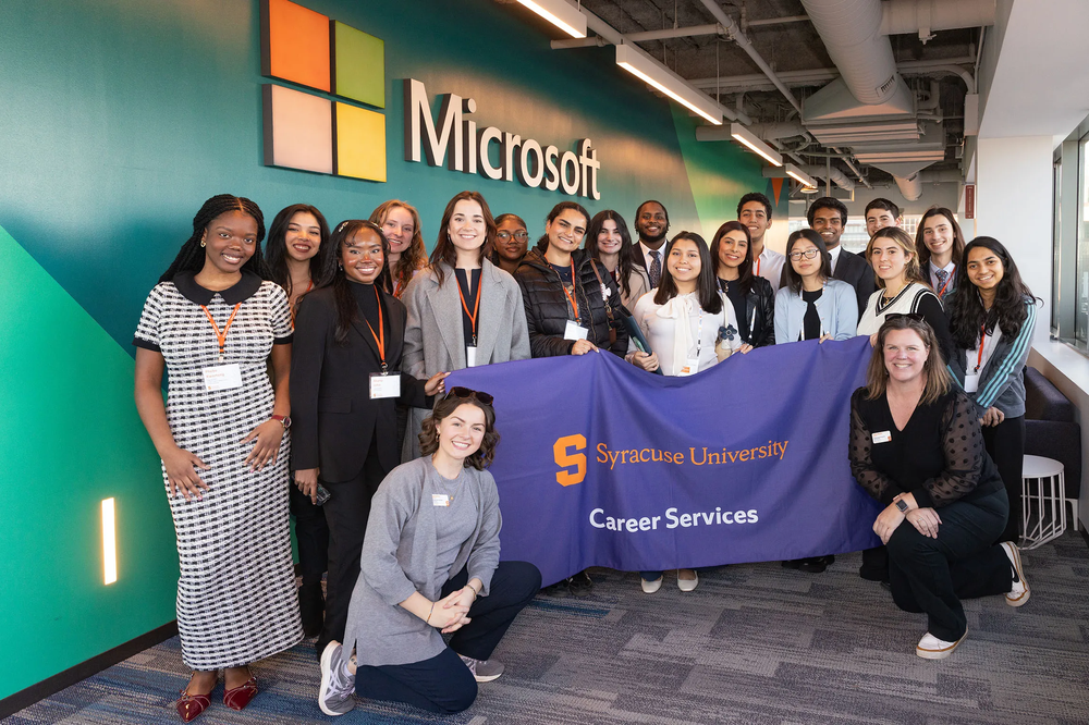 Group of people holding a Syracuse flag with a Microsoft sign in the background.