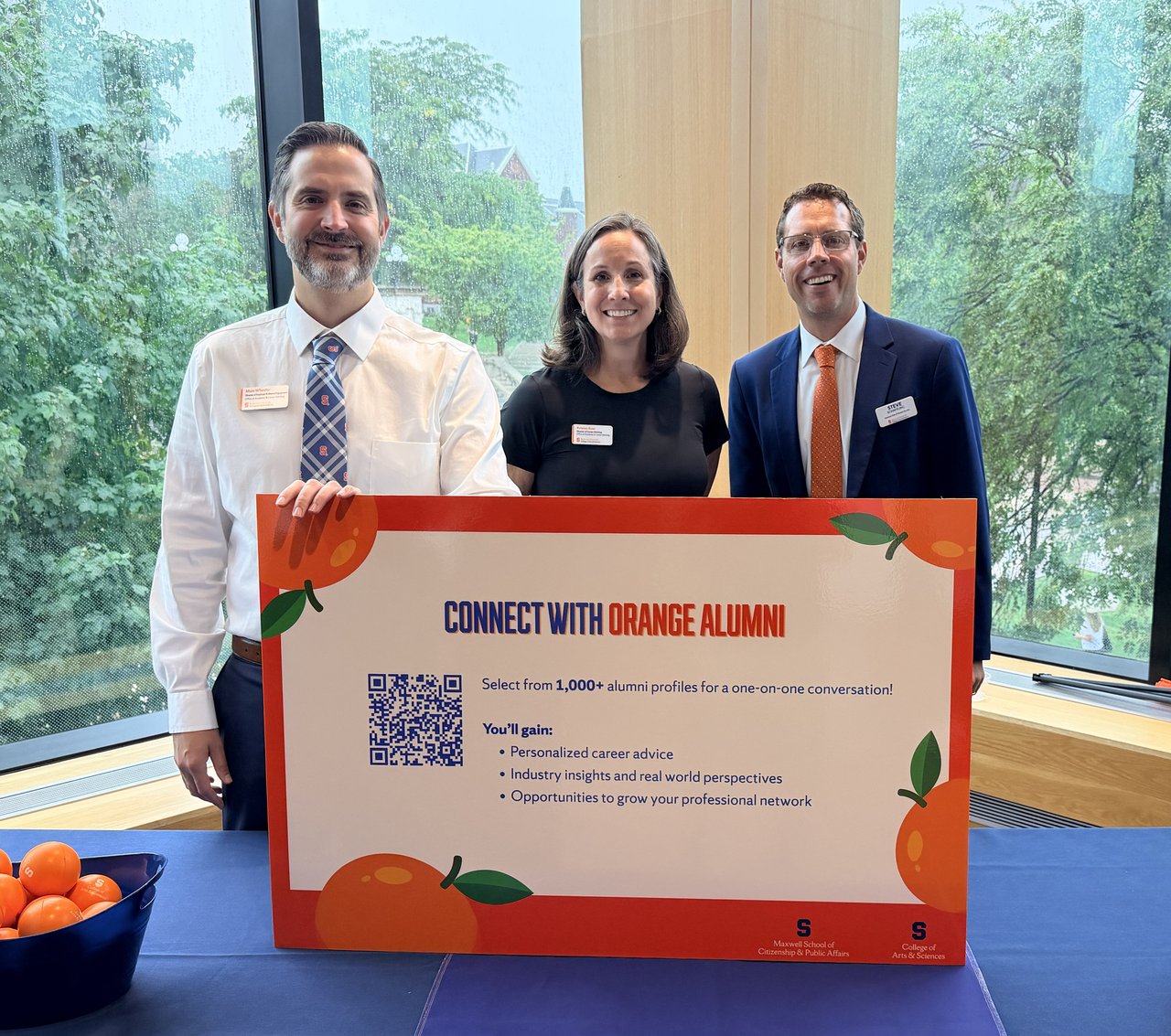 Three smiling adults stand behind a large poster that reads "CONNECT WITH ORANGE ALUMNI," promoting alumni networking; they pose indoors by a rain-speckled window with trees visible outside.