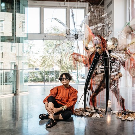 Rina Banerjee seated next to artwork.