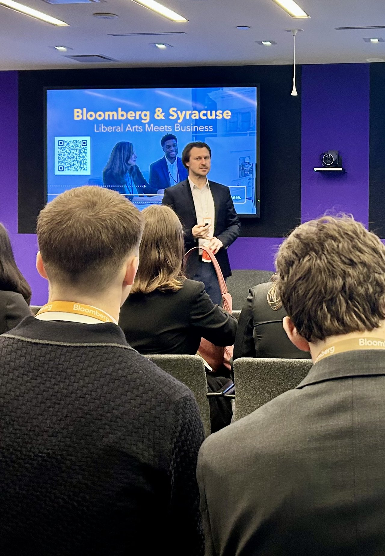 A speaker stands at the front of a conference room before an audience of students wearing Bloomberg lanyards, with a large screen behind him displaying the text 'Bloomberg & Syracuse: Liberal Arts Meets Business.'