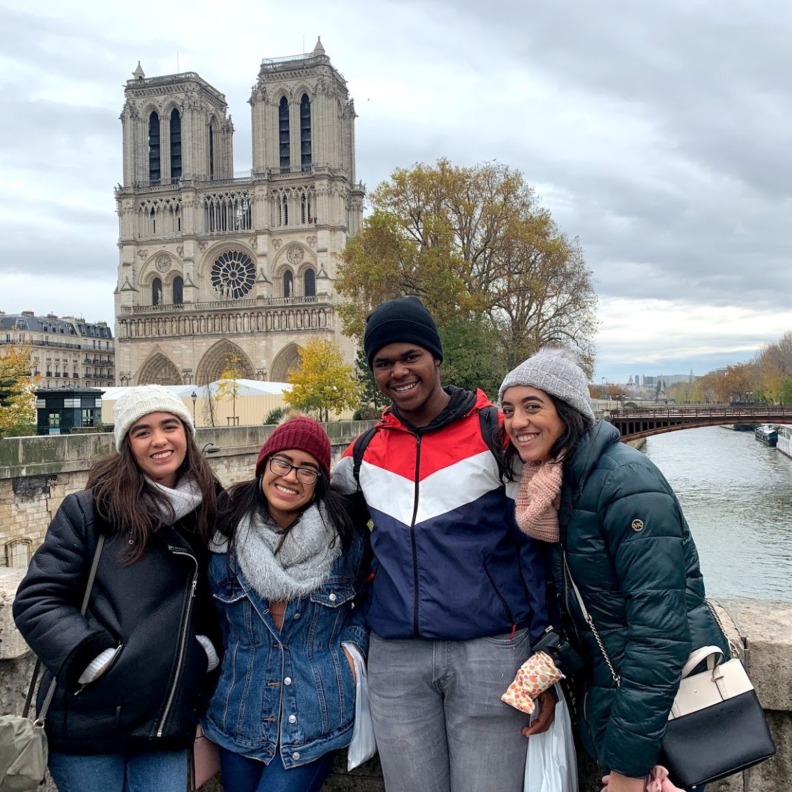 Four smiling young people in winter coats and hats pose together on a bridge along the Seine River, with the Notre-Dame Cathedral and autumn trees visible in the background.