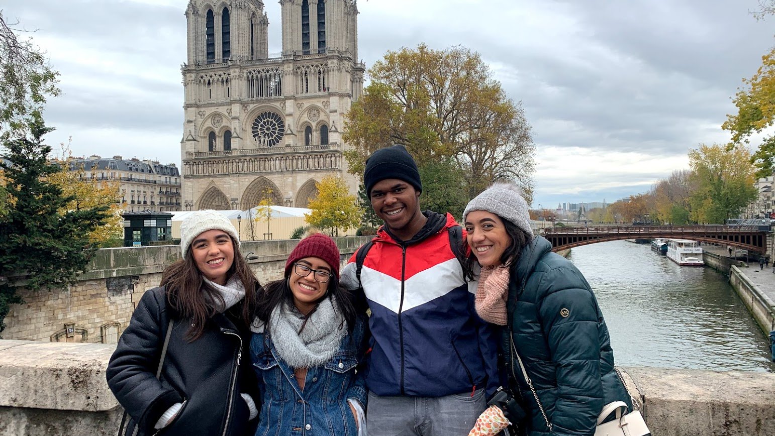 Four smiling young people in winter coats and hats pose together on a bridge along the Seine River, with the Notre-Dame Cathedral and autumn trees visible in the background.