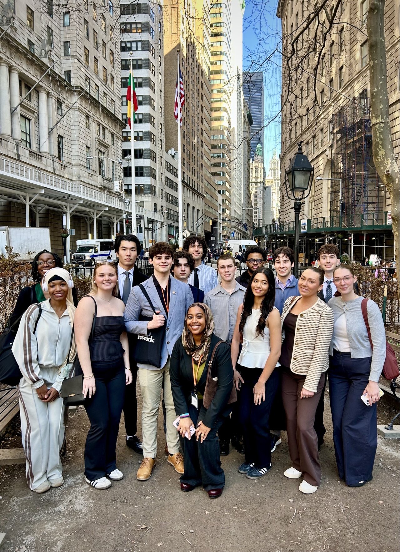 A diverse group of approximately fifteen young adults pose together on a sidewalk in New York City's Financial District, with tall buildings and an American flag visible in the background.