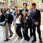 A diverse group of eight professionally dressed young adults pose together on a busy city sidewalk, smiling and making peace signs at the camera. Several are wearing lanyards, suggesting attendance at a professional or academic event.