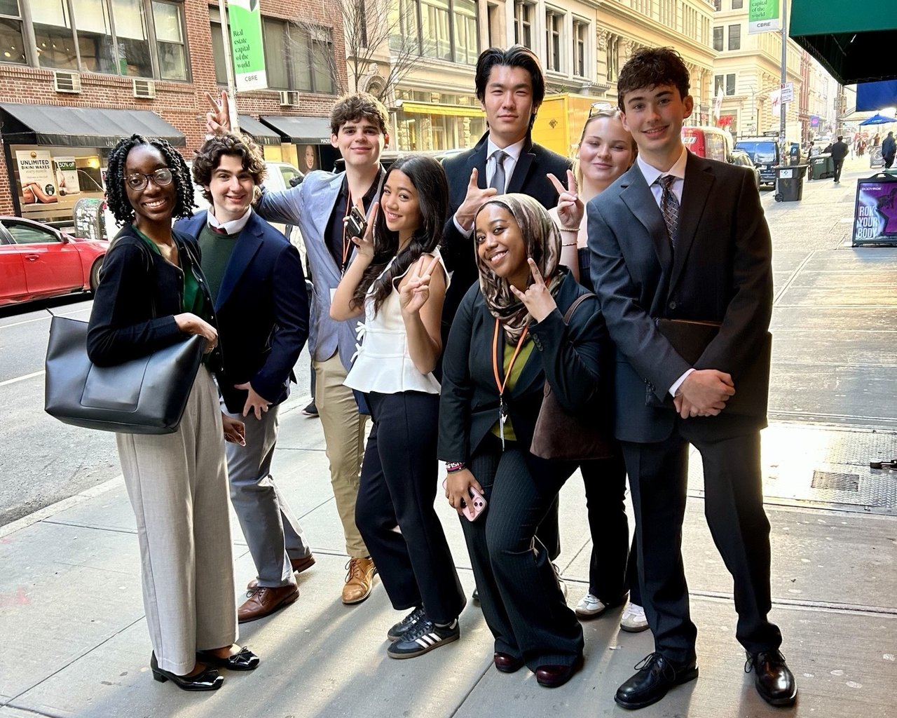 A diverse group of eight professionally dressed young adults pose together on a busy city sidewalk, smiling and making peace signs at the camera. Several are wearing lanyards, suggesting attendance at a professional or academic event.