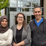 Three people standing together outside with a building in the background.