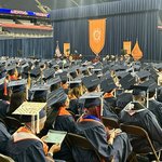 Rows of graduates in caps and gowns sit facing the stage during Convocation, with an orange university banner hanging at the front of the arena.
