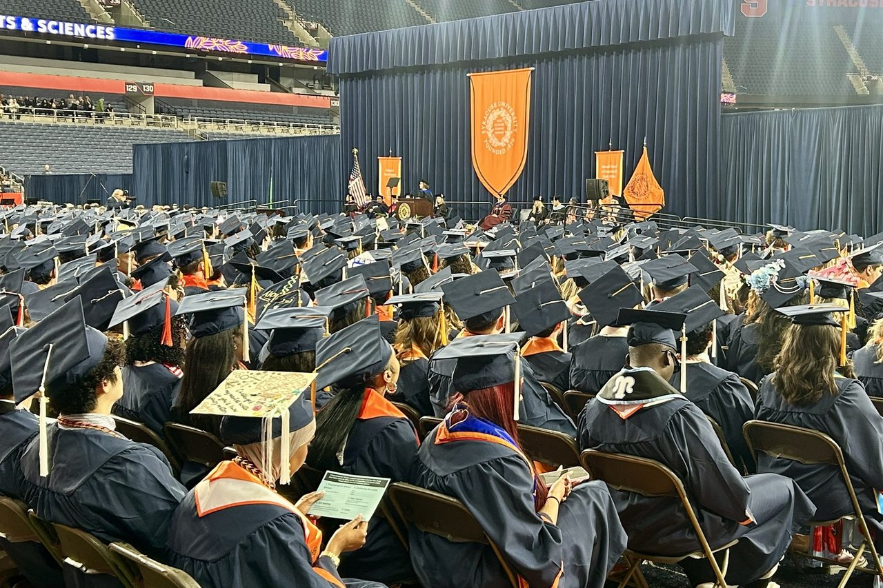 Rows of graduates in caps and gowns sit facing the stage during Convocation, with an orange university banner hanging at the front of the arena.