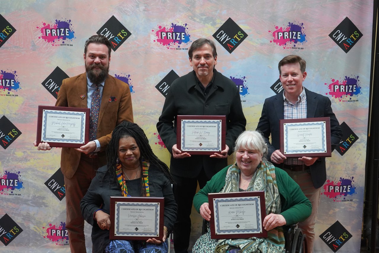 Three people standing and two sitting in front of a CNY Arts Syracuse Prize banner.