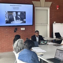 A speaker leads a workshop around a conference table, with a presentation slide titled 'Dalits and Anti-Caste Epistemology' by Dr. Chinnaiah Jangam of Carleton University displayed on the screen behind him.