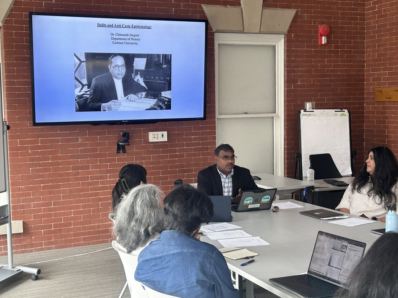 A speaker leads a workshop around a conference table, with a presentation slide titled 'Dalits and Anti-Caste Epistemology' by Dr. Chinnaiah Jangam of Carleton University displayed on the screen behind him.