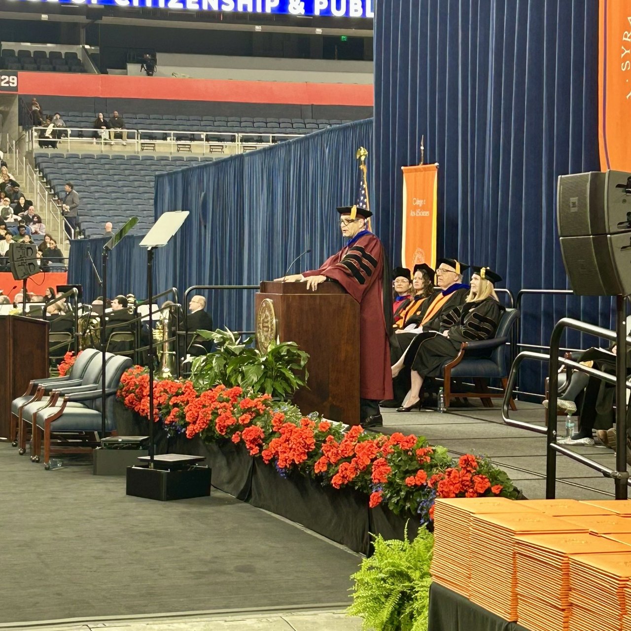 A speaker in maroon doctoral regalia addresses an audience from a podium on stage, with seated faculty in academic robes and a row of red flowers lining the front of the stage.