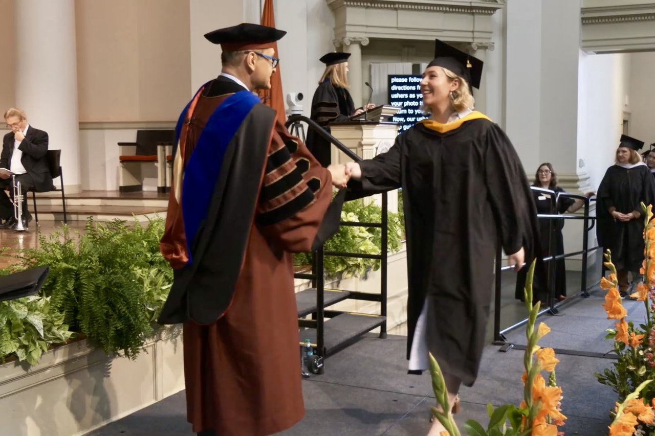 A graduate in cap and gown shakes hands with a faculty member in doctoral regalia on stage during a commencement ceremony, with floral arrangements decorating the front of the stage.
