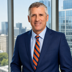A man wearing a suit and orange and blue tie stands near a window.