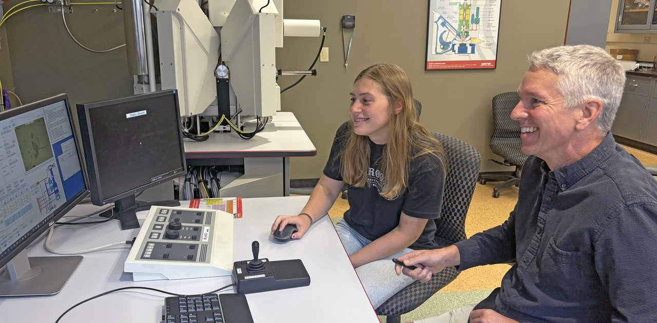 Two people seated at a computer operating an electron micriscope.