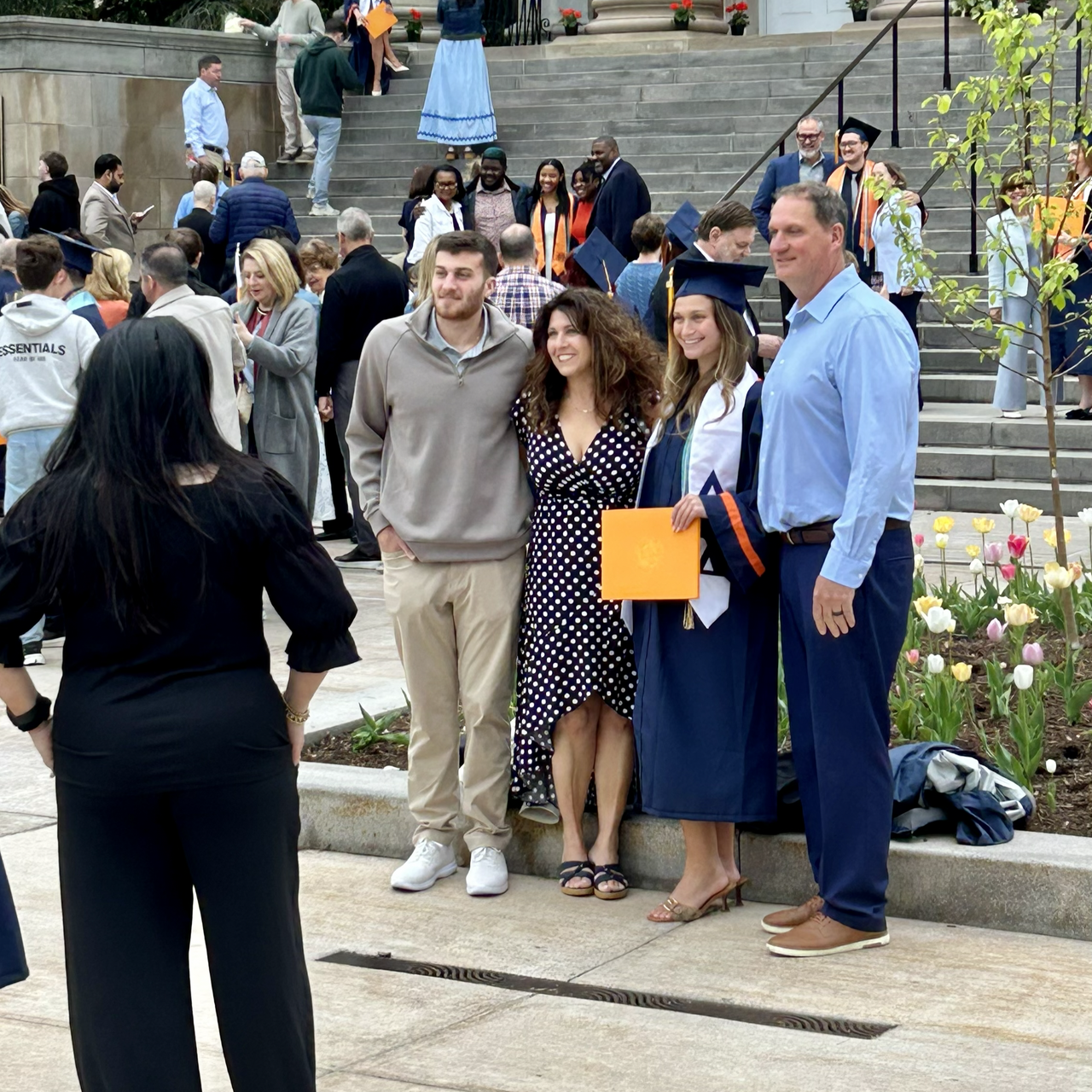 A graduate in cap and gown poses for a family photo while holding an orange Syracuse University diploma folder, with stone steps and tulips visible behind them.