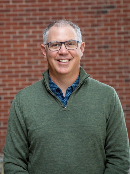 A portrait of Greg Hoke in front of a brick wall.