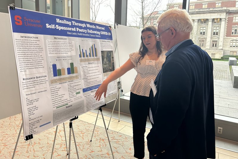 A student gestures toward a Syracuse University research poster titled 'Healing Through Words: Nurses' Self-Sponsored Poetry Following COVID-19' while presenting to an attendee.