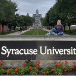 A woman in a Syracuse sweatshirt sits atop the stone wall behind the university’s illuminated entrance sign, with the Hall of Languages and tree-lined promenade visible in the background at dusk, geraniums in the foreground.