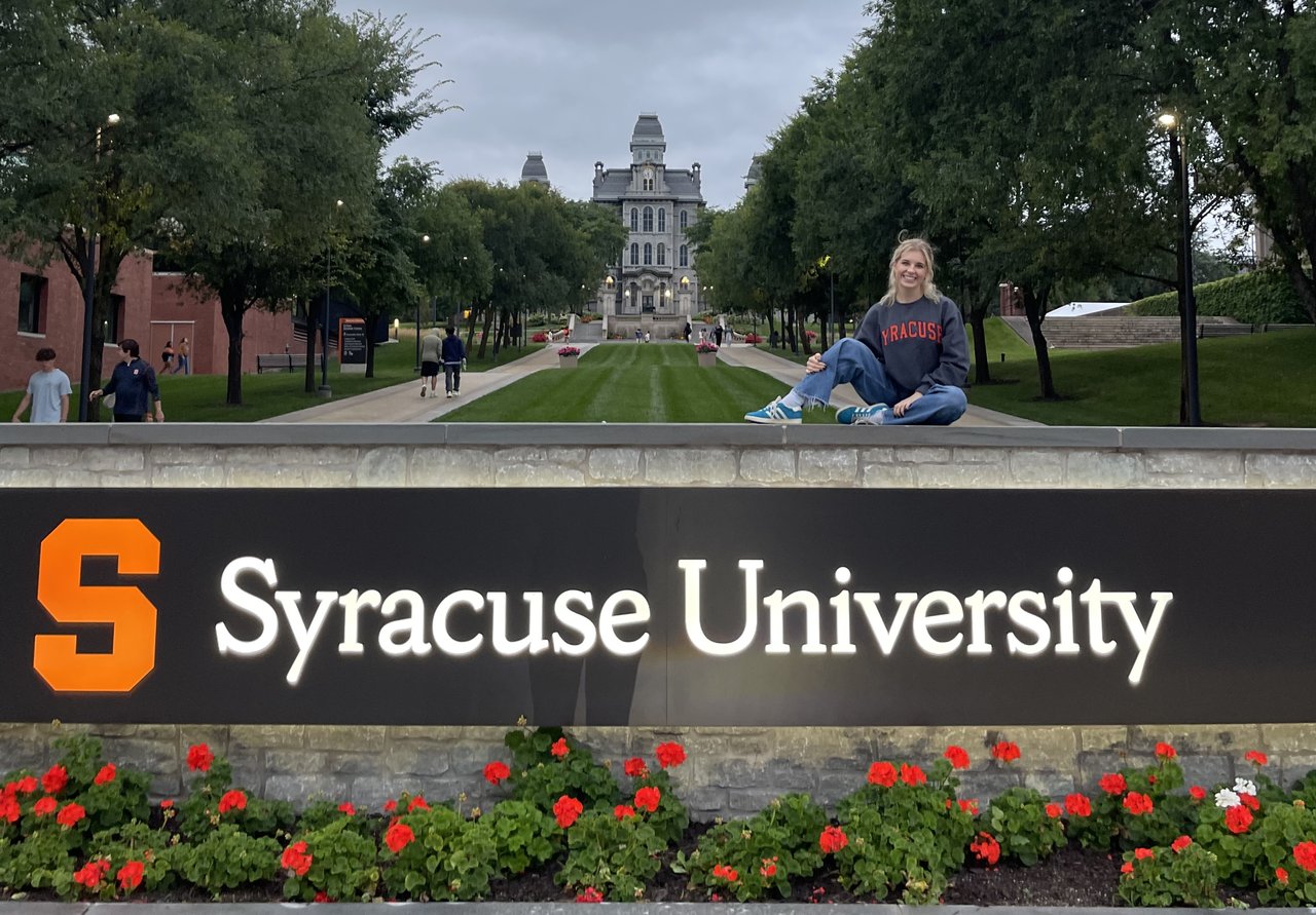 A woman in a Syracuse sweatshirt sits atop the stone wall behind the university’s illuminated entrance sign, with the Hall of Languages and tree-lined promenade visible in the background at dusk, geraniums in the foreground.