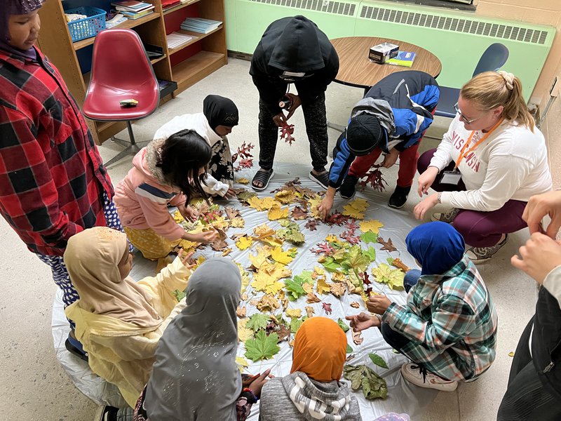 In a classroom, group of kids and a woman surround a mat with leaves of different colors.