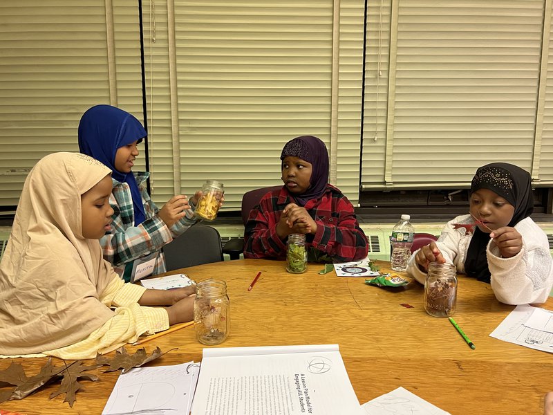 Four girls sitting around a table holding jars with leaves of different colors and talking.