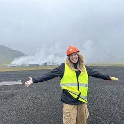 A student, Kenna Cummings, poses in front of the Hellisheidi Geothermal Power Plant in Iceland wearing a reflective vest.