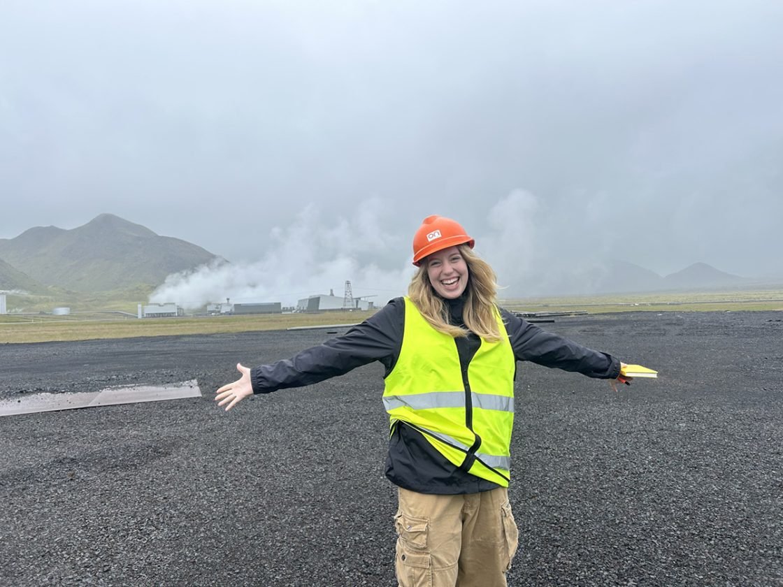 A student, Kenna Cummings, poses in front of the Hellisheidi Geothermal Power Plant in Iceland wearing a reflective vest.
