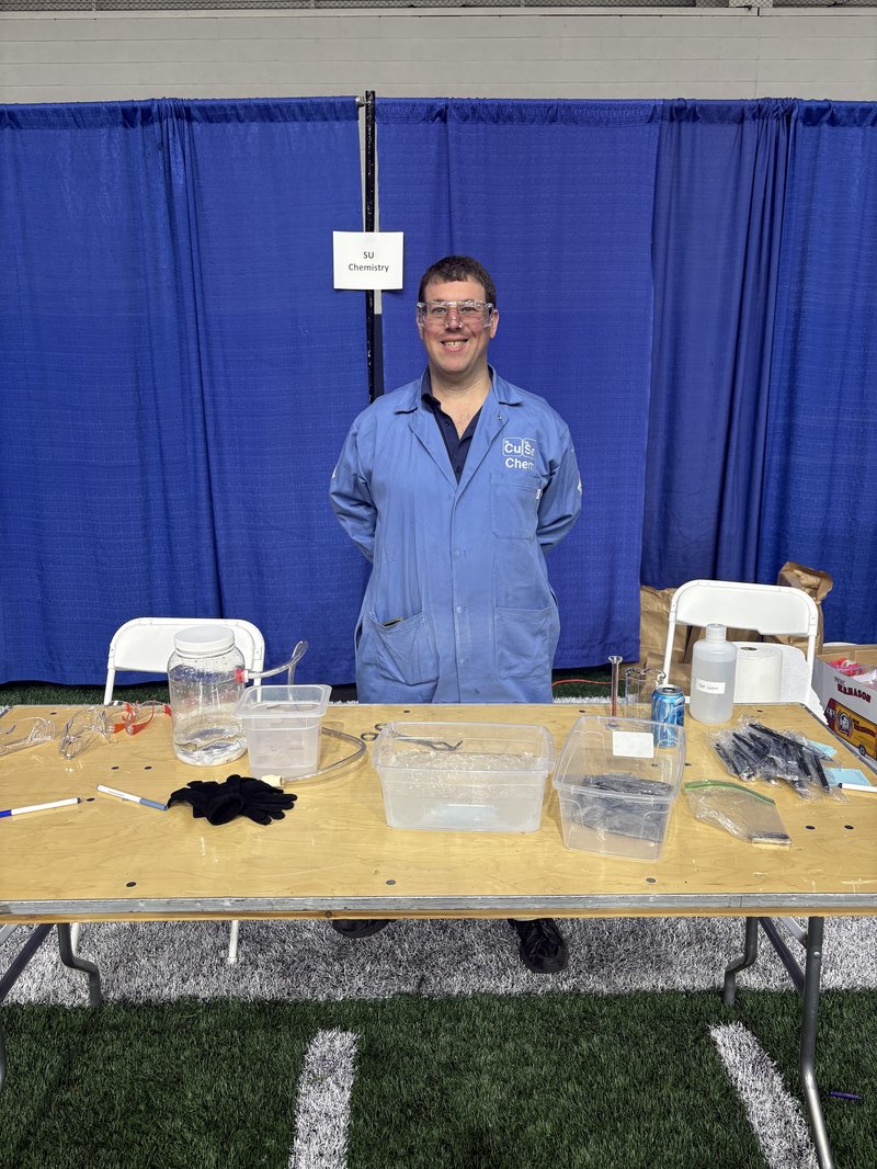 A person wearing safety goggles and a blue lab coat stands behind a table of chemistry demonstration supplies at an outreach event, with an 'SU Chemistry' sign posted on a blue curtain behind him.