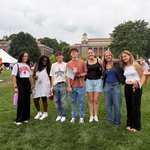 Seven students pose together on the quad during a campus event, with cornhole boards, orange and navy balloon columns, and Carnegie Library visible in the background.