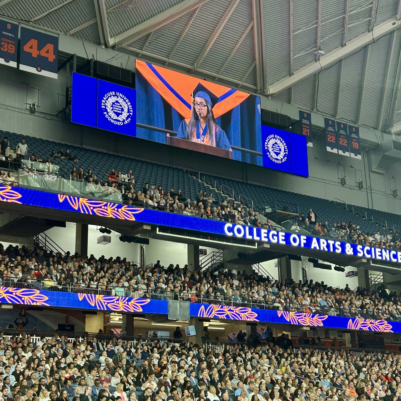 A student in graduation regalia speaks at a podium, shown on a large jumbotron screen above a packed arena audience at the College of Arts & Sciences convocation.