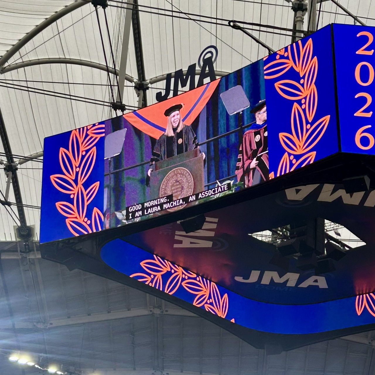 A large jumbotron screen inside the JMA Dome shows a faculty speaker in academic regalia at a podium, with closed captions reading 'Good morning. I am Laura Machia, an associate.'