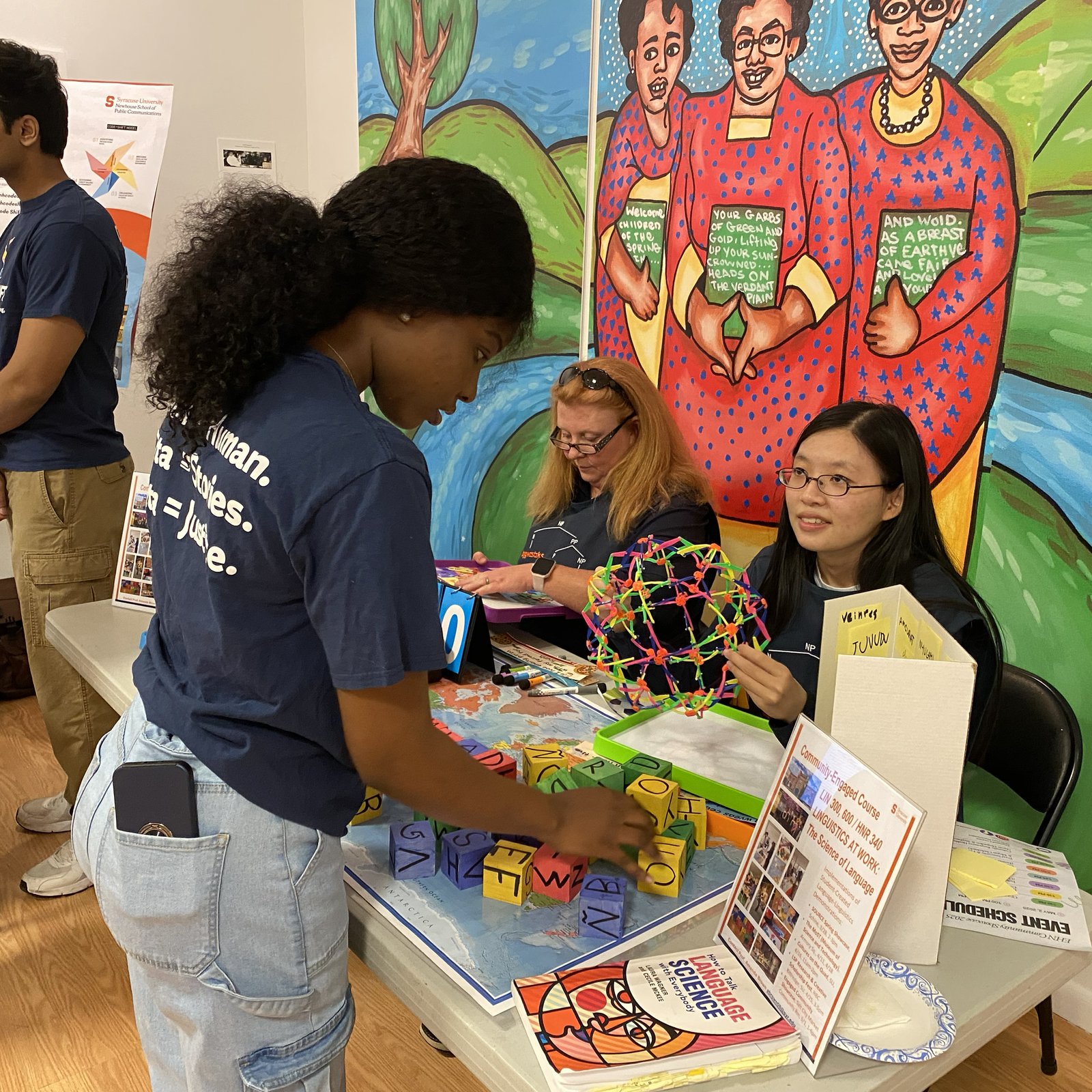 A student interacts with two representatives at a table promoting a "Linguistics at Work" community-engaged course, which is decorated with colorful letter blocks and a geometric model, set against a vibrant painted mural backdrop.