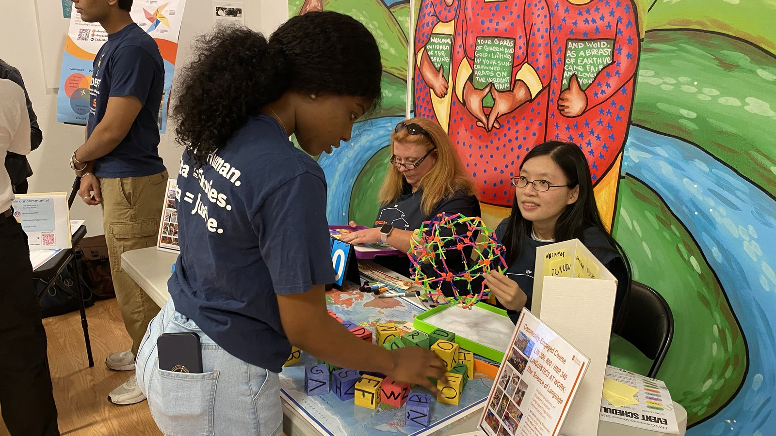 A student interacts with two representatives at a table promoting a "Linguistics at Work" community-engaged course, which is decorated with colorful letter blocks and a geometric model, set against a vibrant painted mural backdrop.