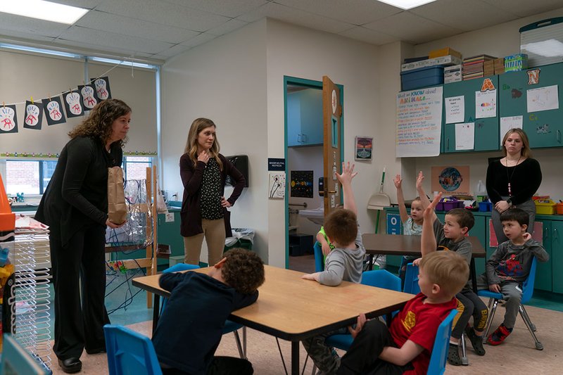 Adults in a classroom with children raising their hands.