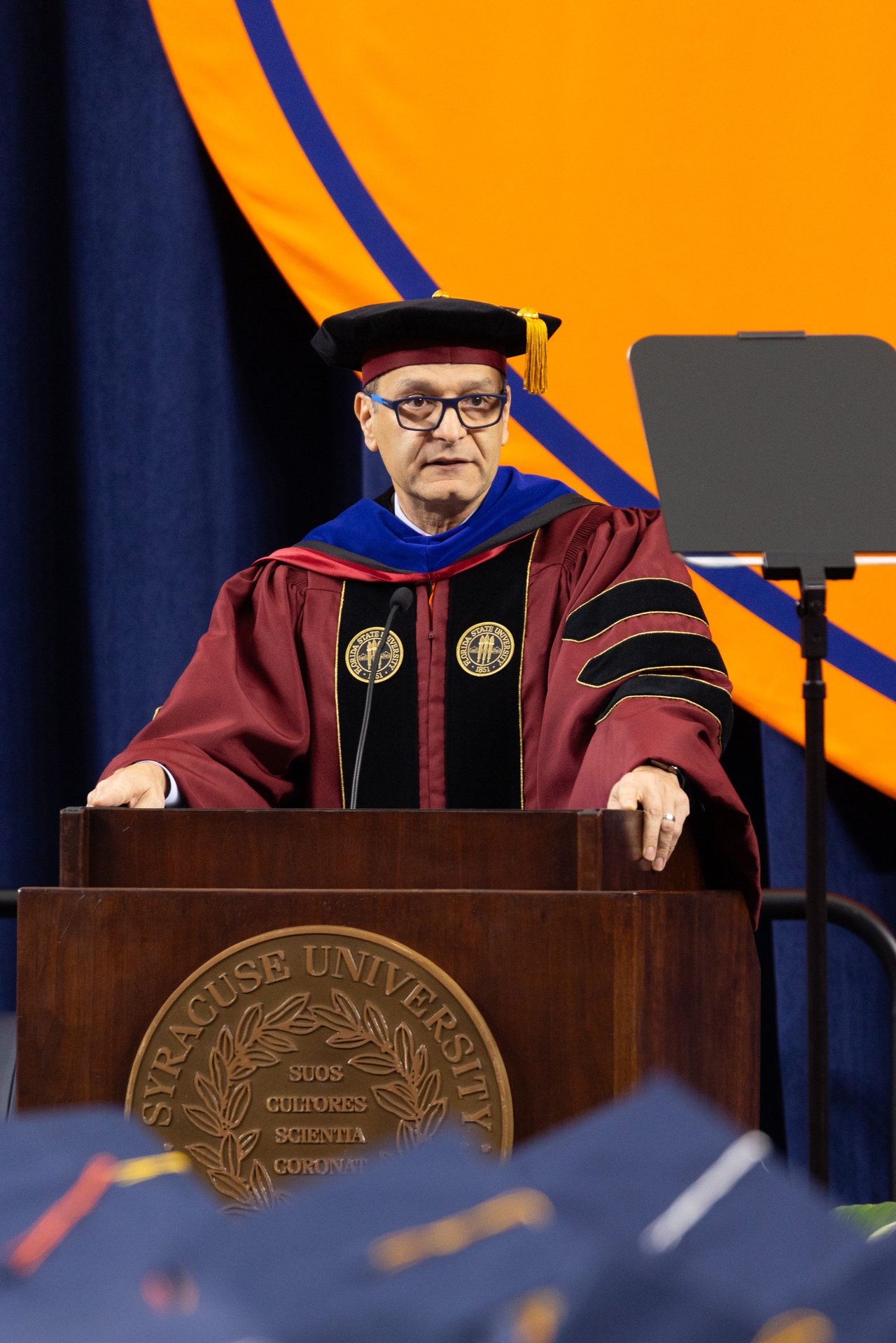 A speaker in maroon doctoral regalia addresses an audience from a podium on stage, with seated faculty in academic robes and a row of red flowers lining the front of the stage.